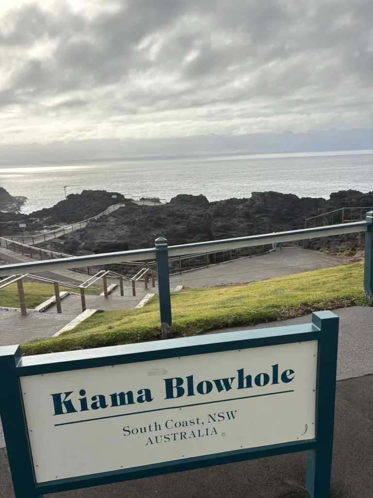 A scenic view of the Kiama Blowhole lookout in New South Wales, Australia, with rocky cliffs, pathways, and the ocean under a cloudy sky.