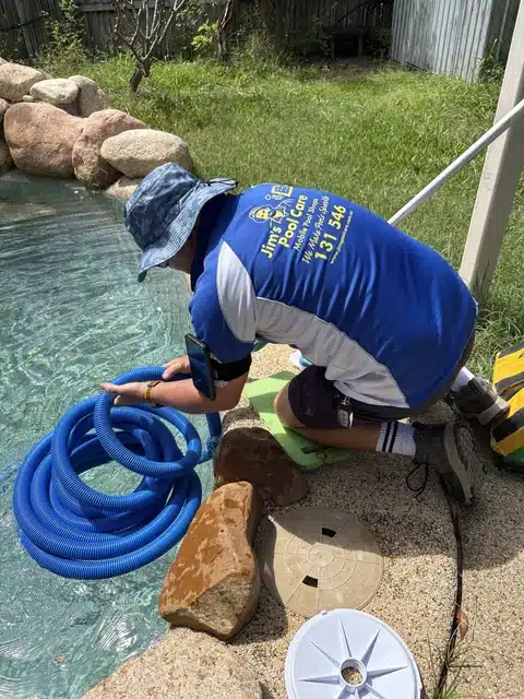 Jim’s Pool Care Townsville technician in branded uniform coiling a vacuum hose beside a rock-edged swimming pool