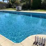 Pool water balancing test kit and chemical reagent bottles on the coping of a clear blue backyard swimming pool