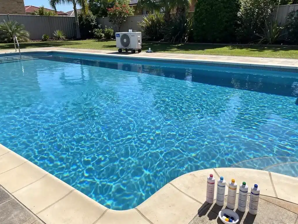 Pool water balancing test kit and chemical reagent bottles on the coping of a clear blue backyard swimming pool