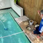 Jim’s Pool Care technician cleaning a pool skimmer basket while a suction cleaner runs in a clear backyard pool